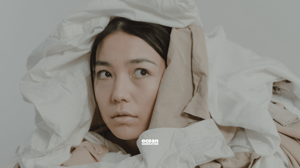 Asian woman surrounded by a pile of clothing. Just her face showing amongst all the clothing and textiles. This photo represents the overconsumption in the fashion industry.