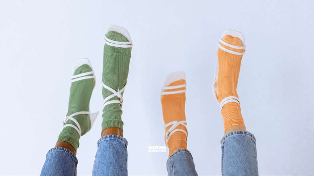 Two sets of socked feet are up in the air. One pair of feet is wearing green socks and the other is wearing mustard yellow socks. They are both wearing white strappy high heels and blue jeans. 