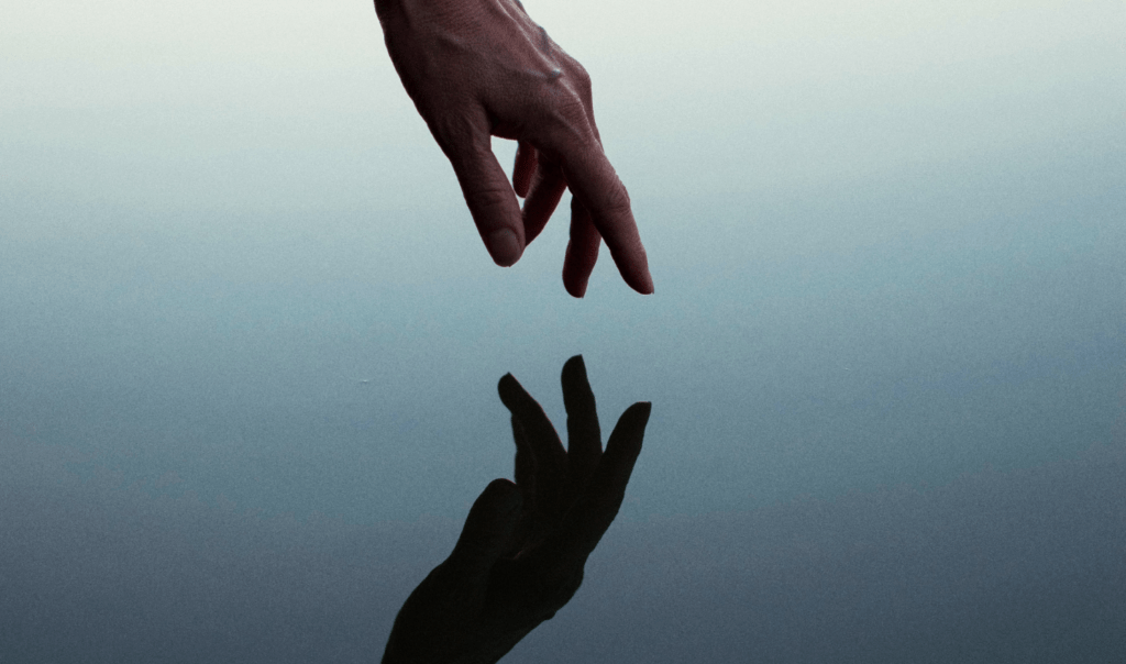 A hand reaching out above a body of water. The hand's reflection looms below. Shared by Ocean Generation.