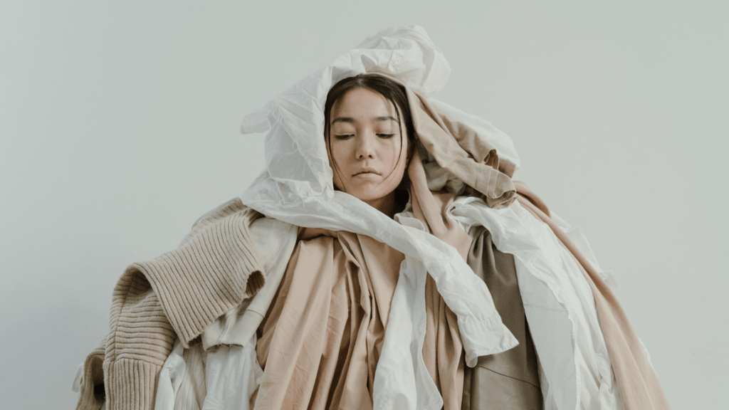 Asian woman surrounded by neutral coloured clothing, emphasising our overconsumption mentality. Ocean Generation is sharing 5 strategies to become a more conscious consumer in this article.