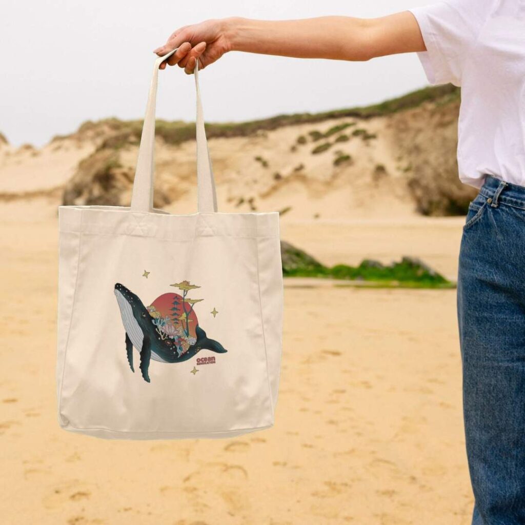 Woman on the beach, with an Ocean-friendly beach bag sold by Ocean Generation. Whale beach bag. Ocean Generation's clothing products are all sustainable, using organic cotton and 100% renewable energy to manufacture the items.