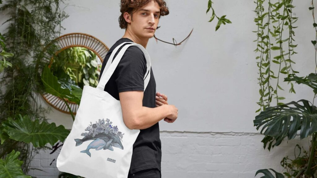 Organic cotton whale tote bag, perfect for Ocean lovers. Man carrying a tote bag with a whale design on it. Ocean-friendly clothing is sold by Ocean Generation.