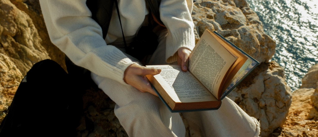 Man reading a book on a rocky coast with waves crashing nearby.